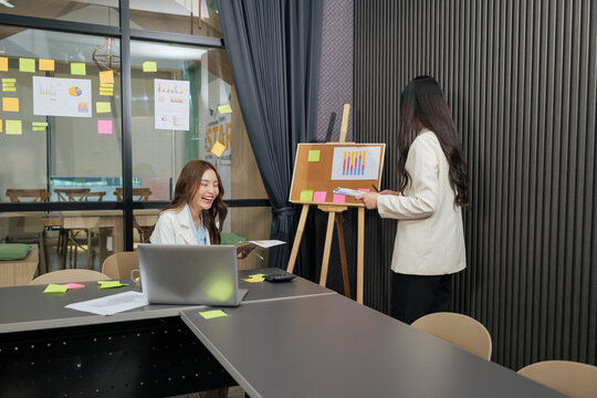 Business woman, busy team members meet for project work, planning meeting, smiling woman with laptop reviews data, colleague presents strategy on cork board with charts, modern office collaboration - Powered by Adobe