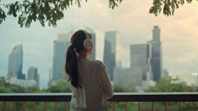 Asian woman listening to music with headphones while looking at the city skyline, relaxing on balcony, urban lifestyle, peaceful morning mood, modern city living and mindfulness moment