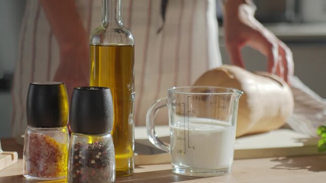 Woman placing measuring cup with cream on kitchen table while preparing food. Cooking process with milk, olive oil, salt, pepper and squash at home kitchen