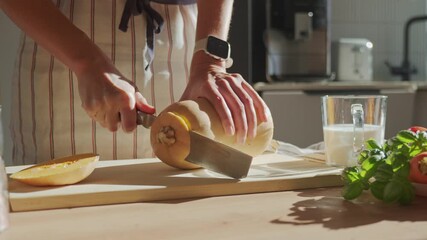 Person in apron cutting butternut squash on wooden board in kitchen. Close-up of woman slicing fresh pumpkin with fresh vegetables and herbs on table. Concept of healthy eating and home cooking