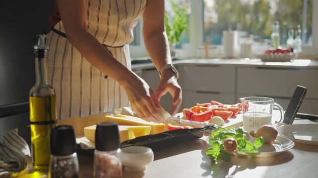 Woman in striped apron placing butternut squash or pumpkin on baking tray in bright kitchen with fresh vegetables and ingredients. Process of cooking at home and healthy food preparation