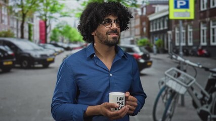 Man with curly hair wearing blue shirt holding a white coffee mug with both hands and smiling while looking up on a city street; calm reflection.