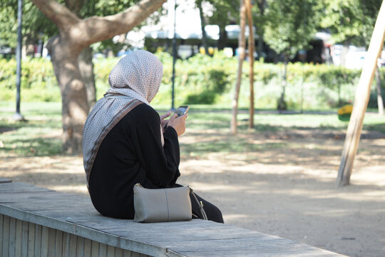Woman in hijab sitting on a bench using her phone in park - Powered by Adobe