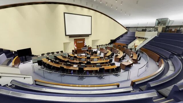 An interior wide shot of the iconic, unoccupied Toronto City Council Chamber in City Hall. A large presentation screen is visible above the mayor's central seat