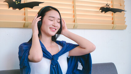Young woman enjoying music with headphones, wearing a blue cape, relaxing indoors, surrounded by Halloween decorations, cheerful mood and joyful expression.