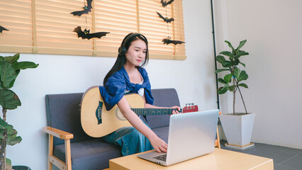 Young Woman Playing Acoustic Guitar While Using Laptop in Cozy Indoor Setting Surrounded by Halloween Decorations and Potted Plants
