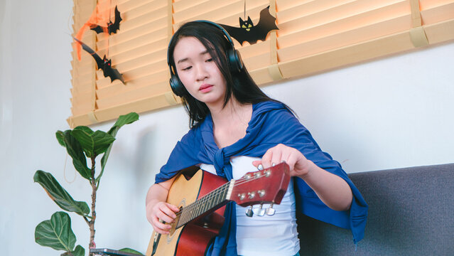 Young woman playing acoustic guitar indoors with headphones wearing casual attire and festive decorations for Halloween in cozy home setting
