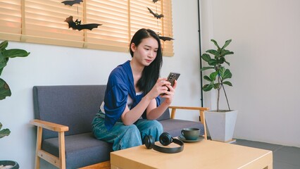 Young woman using smartphone in cozy living room with plants and Halloween decorations, enjoying personal time and relaxation in modern interior setting