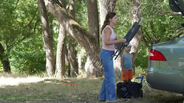 A green folding chair is packed by the mother near the open car trunk. The waiting boy stands on the dry grass as they finish their forest picnic.