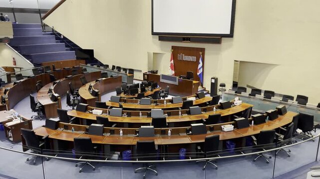 An elevated, side-angle view from the public gallery looking down at the unoccupied Toronto City Council Chamber. The presentation screen and flags are visible.