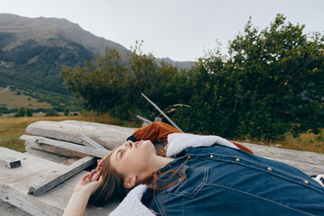 Fototapeta premium Woman lying on wooden bench in nature with mountains background, relaxed portrait in denim jacket outdoors, cozy layers and peaceful relaxation during countryside escape.