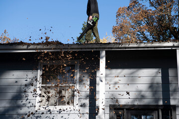 Man walks along roof line to remove leaves and debris from the gutter of a suburban home on a fall day. © Jessie