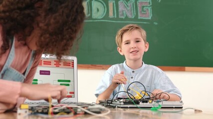 Caucasian energetic boy fixing main board and looking at camera while american student working together to inspect electronic machine by using color wires at table with laptop display code. Pedagogy. - Powered by Adobe