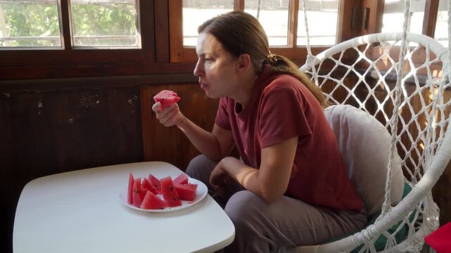 A woman in a hanging macrame chair eats a slice of red watermelon. She holds the fresh fruit while sitting at a white table on a sunny wooden porch.