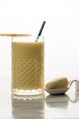 Banana milkshake drink in a patterned glass with a black straw and a wafer, accompanied by a piece of banana on a white spoon