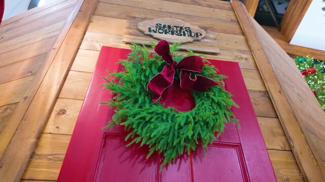 Victoria, BC, Canada - November 25, 2025: Green wreath with a red bow adorns a red door under Santa's Workshop sign. Slow motion