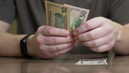 Male hands carefully counting united states dollar banknotes at a wooden table, calculating income, savings, or payment for a purchase before placing the cash down and clasping his hands