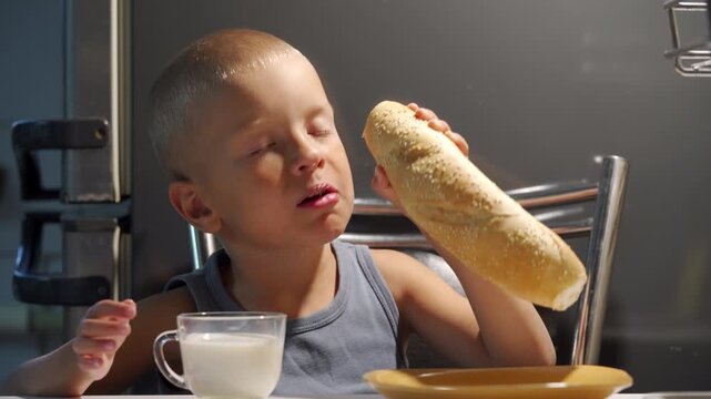 Holding a sesame bread loaf, the hungry boy opens his mouth in anticipation. Near a glass milk cup, the child sits at the kitchen table ready to eat.