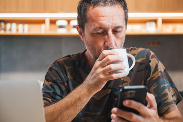 Procrastination concept, mature adult caucasian male wearing camouflage army shirt drinking coffee and using mobile phone at home