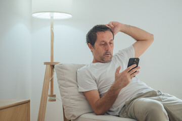 Fototapeta premium Laid back mature adult male bored and tired procrastinating while looking at social media content on smartphone device in cozy living room chair next to floor lamp
