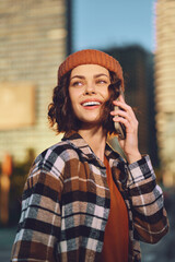 Woman on phone smiles in city portrait wearing beanie and plaid jacket, outdoor sunlight casting...