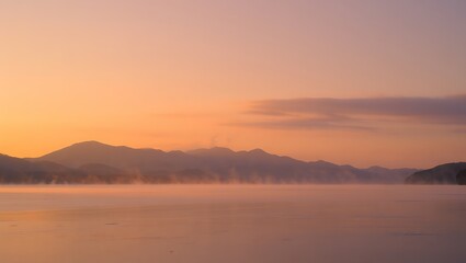 Serene mountain range silhouetted against a soft peach and orange sunrise sky over a calm misty lake