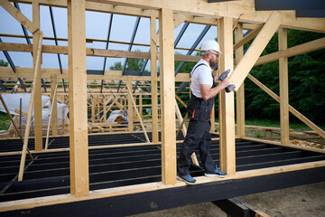 Construction worker using an electric screwdriver while building a wooden frame structure. Man in protective helmet, gloves. Concept of construction, carpentry, engineering and building process.