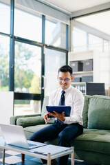Asian businessman sitting on office sofa using smartphone and laptop, smiling while working with documents in a modern workspace.