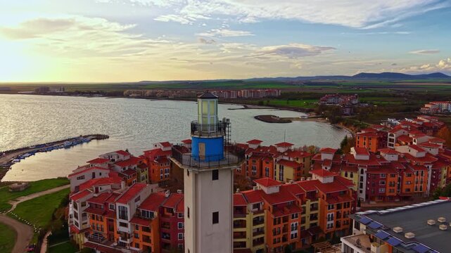 A tall lighthouse overlooks a lively town with red-roofed buildings, set against a backdrop of shimmering water and green fields under a clear sky. The scene radiates warmth and charm.