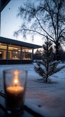 A lit candle in a glass is in the foreground, with a snow-covered pine sapling and a building in the background at dusk.