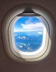 Aerial view of mountainous terrain and cloud-filled sky as seen through an airplane window. Sunlight casts shadows on frame