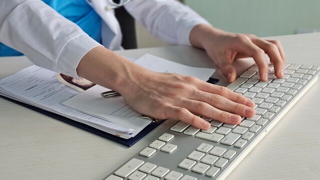 Hands of a medical professional typing on a keyboard while reviewing documents and files