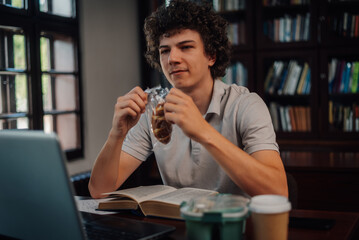 Student eating snack while studying in library with laptop