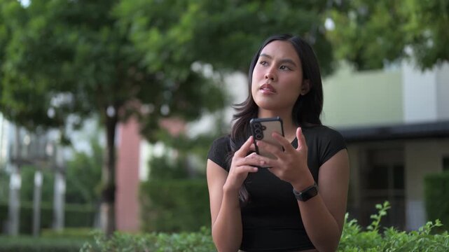 Thoughtful young Asian woman using technology outdoors while sitting in park alone while planning
