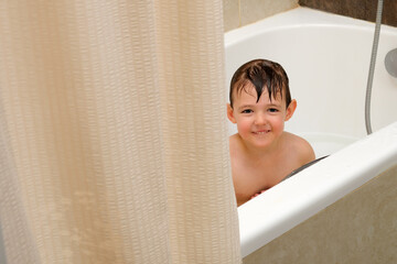Smiling child boy enjoying a fun bath behind a shower curtain in a cozy bathroom, showing...