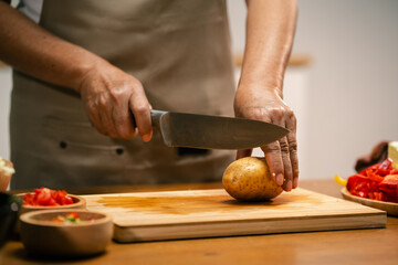 Close-up of Asian man's hands slicing potatoes on a wooden cutting board in the kitchen, preparing for cooking.