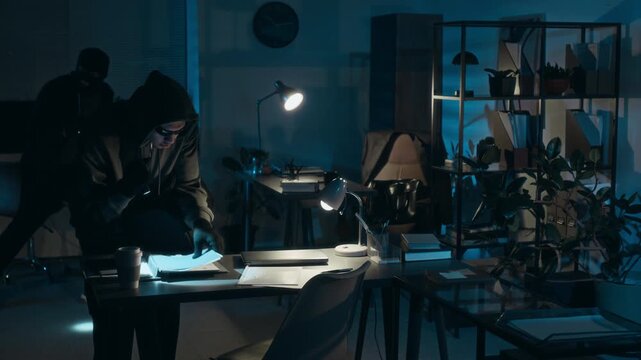 Male criminal examining business papers and stealing laptop from work desk during office burglary with accomplice in background