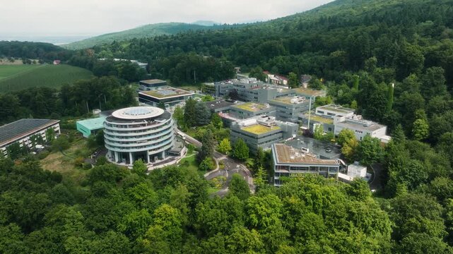 A drone descends slightly while approaching the DKFZ campus, coming from the hillside and fields, revealing the round tower, green roofs, and the forested surroundings