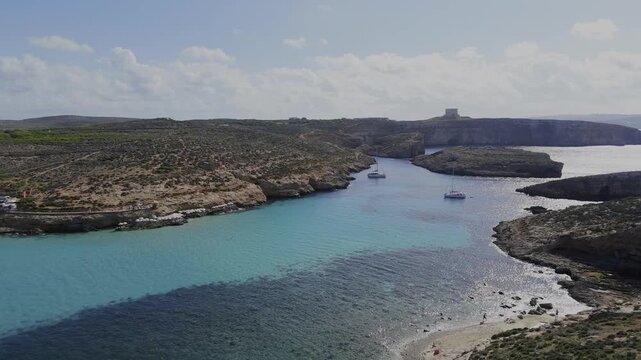 A calm panoramic drone shot over the Blue Lagoon shows Comino, Cominotto Beach and two yachts, with bright sunlight reflecting on clear turquoise Mediterranean water.