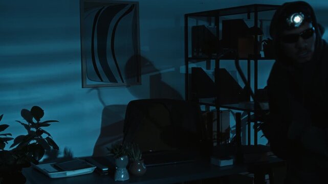 Young male criminal in balaclava holding flashlight and his accomplice with headlamp and crowbar carefully walking around office at night