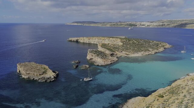 A high drone view over the Blue Lagoon shows Gozo, the open sea, part of Comino and Cominotto Beach with its cave, several yachts and a boat moving across calm turquoise water under sunny skies.