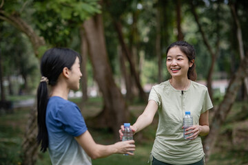 Fototapeta premium A young, happy, and smiling Asian woman gives a bottle of water to her friend after finishing an exercise in the park, surrounded by green trees and a refreshing, calm, and relaxed atmosphere