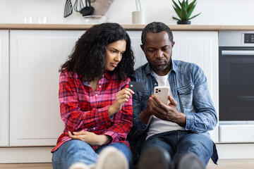 A couple is sitting on the floor, using a credit card and a smartphone to shop online in their kitchen.