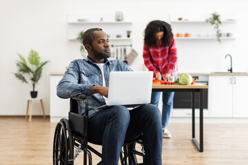 A man in a wheelchair works on a laptop while his partner prepares food in the kitchen.