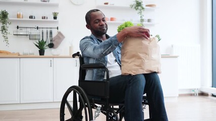 A smiling Black man in a wheelchair holds a bag of fresh groceries in his kitchen, promoting healthy eating.