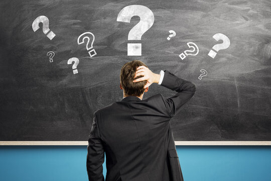 Confused businessman scratching head in front of chalkboard with question marks symbolizing problem-solving and curiosity in education or business.