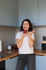 Smiling woman with braces holding a red apple in the kitchen, playful and cheerful moment during a casual home scene