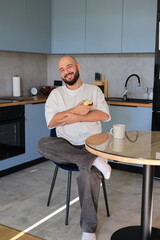 Smiling man holding an apple while sitting in the kitchen, enjoying a relaxed and cheerful moment at home