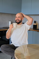 Smiling man holding a cup of coffee, scratching his head while laughing in a relaxed and cheerful home kitchen scene