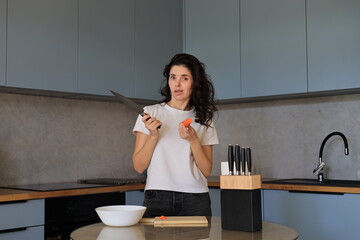 Woman with braces holding a knife and a carrot in the kitchen, smiling playfully while preparing food during a home cooking routine